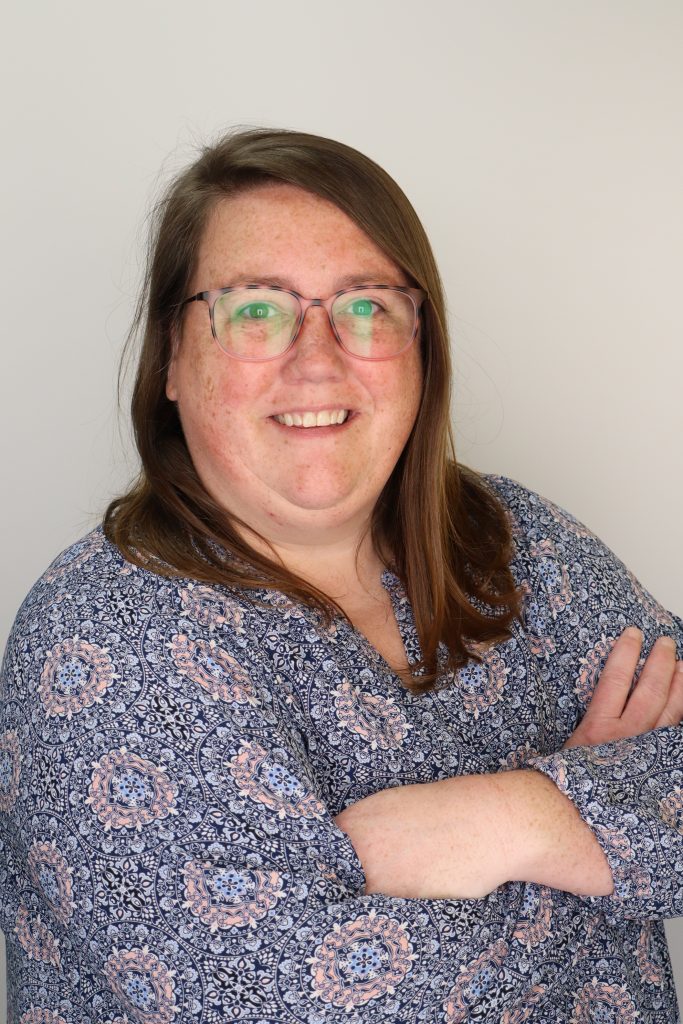 Megan Walters is wearing a blouse with blue and pink geometric pattern. She is wearing glasses and has brown hair just below her shoulders.